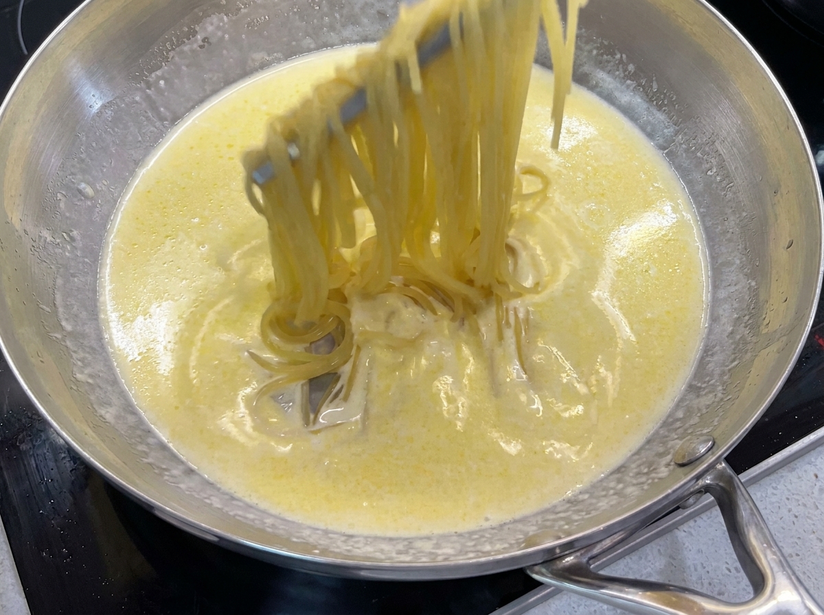 Using metal tongs to lift and toss cooked spaghetti in a bubbling creamy butter sauce inside a pan.