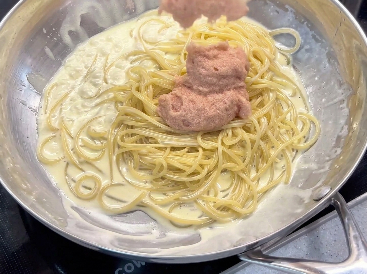 Adding a scoop of pink Mentaiko and mayonnaise paste to the creamy coated spaghetti in a pan.