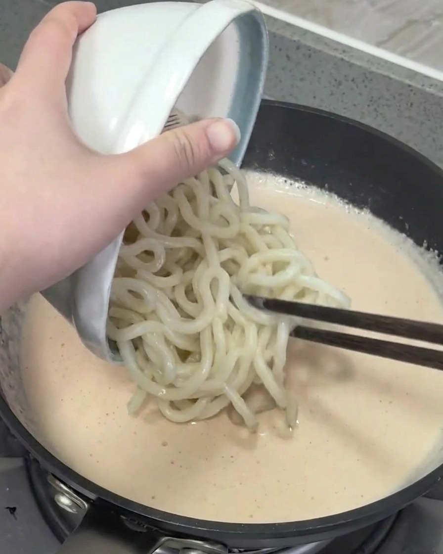 Mixing cooked udon noodles into the mentaiko cream sauce in the pan.