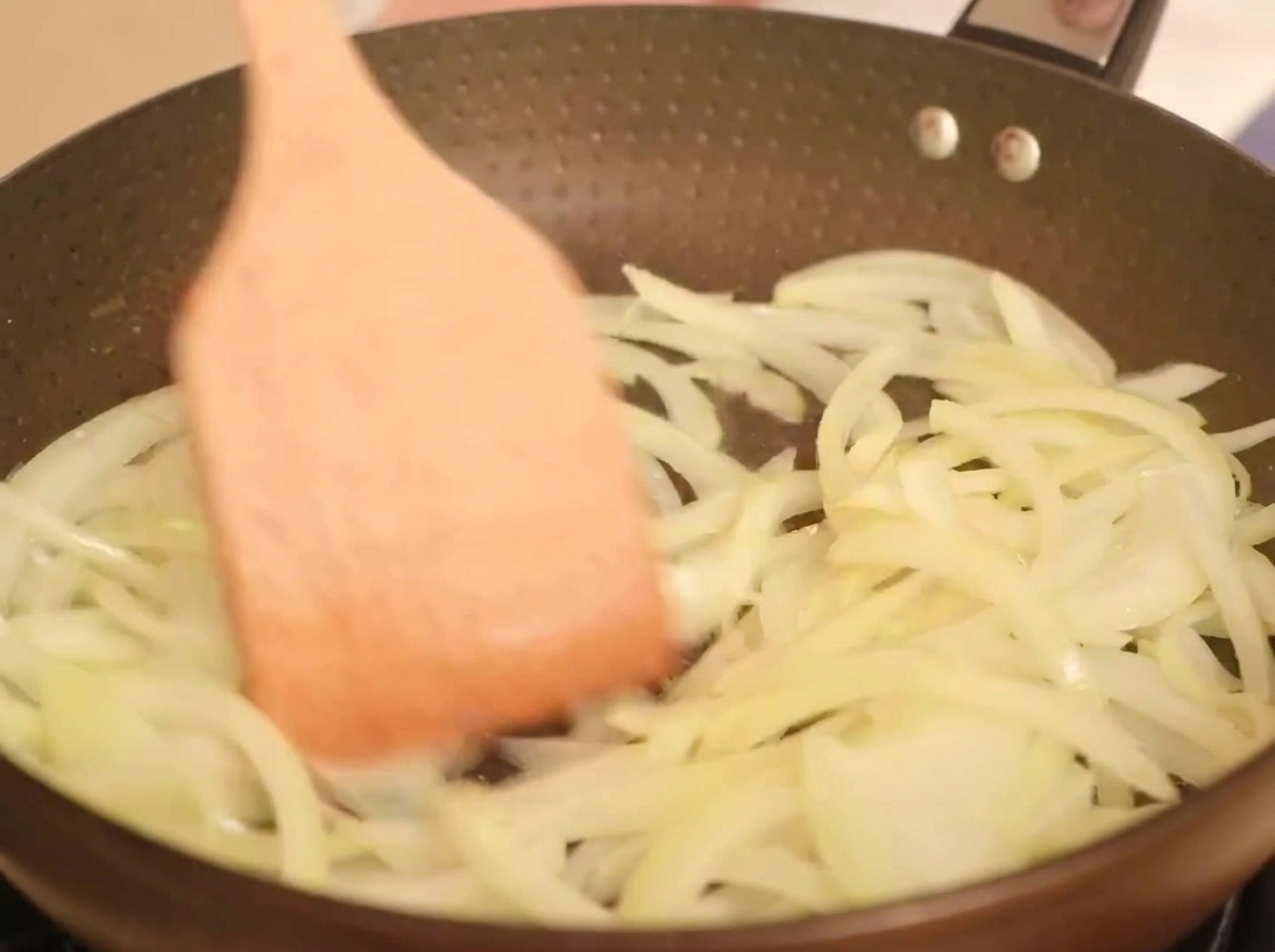 Slices of yellow onion cooking in a hot pan with oil, being tossed with a wooden spatula.
