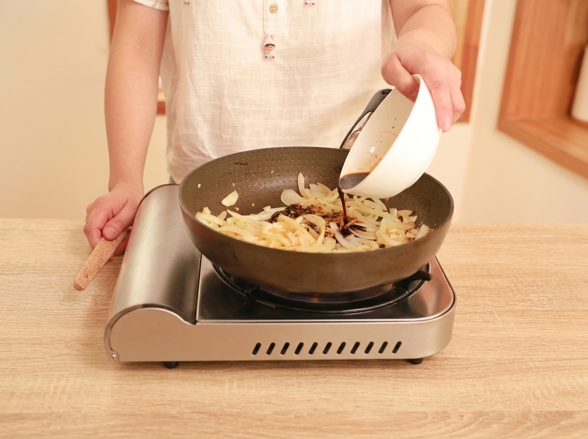 Dark brown sauce being poured from a white bowl into a pan containing sautéed onions, ginger, and garlic.