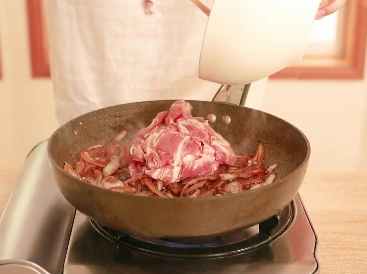 Raw, thinly sliced beef being added to a pan with a dark sauce and softened onions, ready to be stir-fried.
