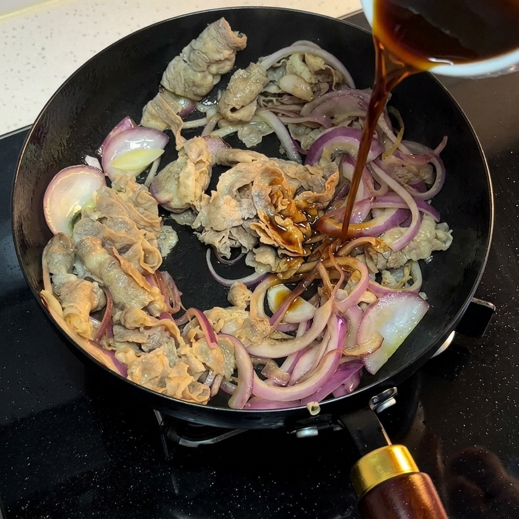 A dark sauce being poured from a small bowl into a pan containing browned beef and onion slices.