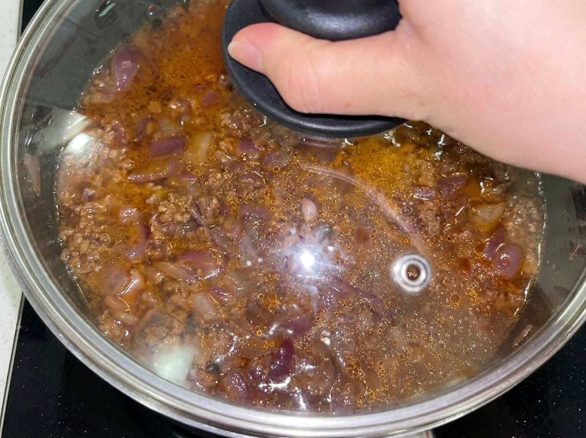 Beef and onion mixture in a pan covered with a glass lid after adding water.