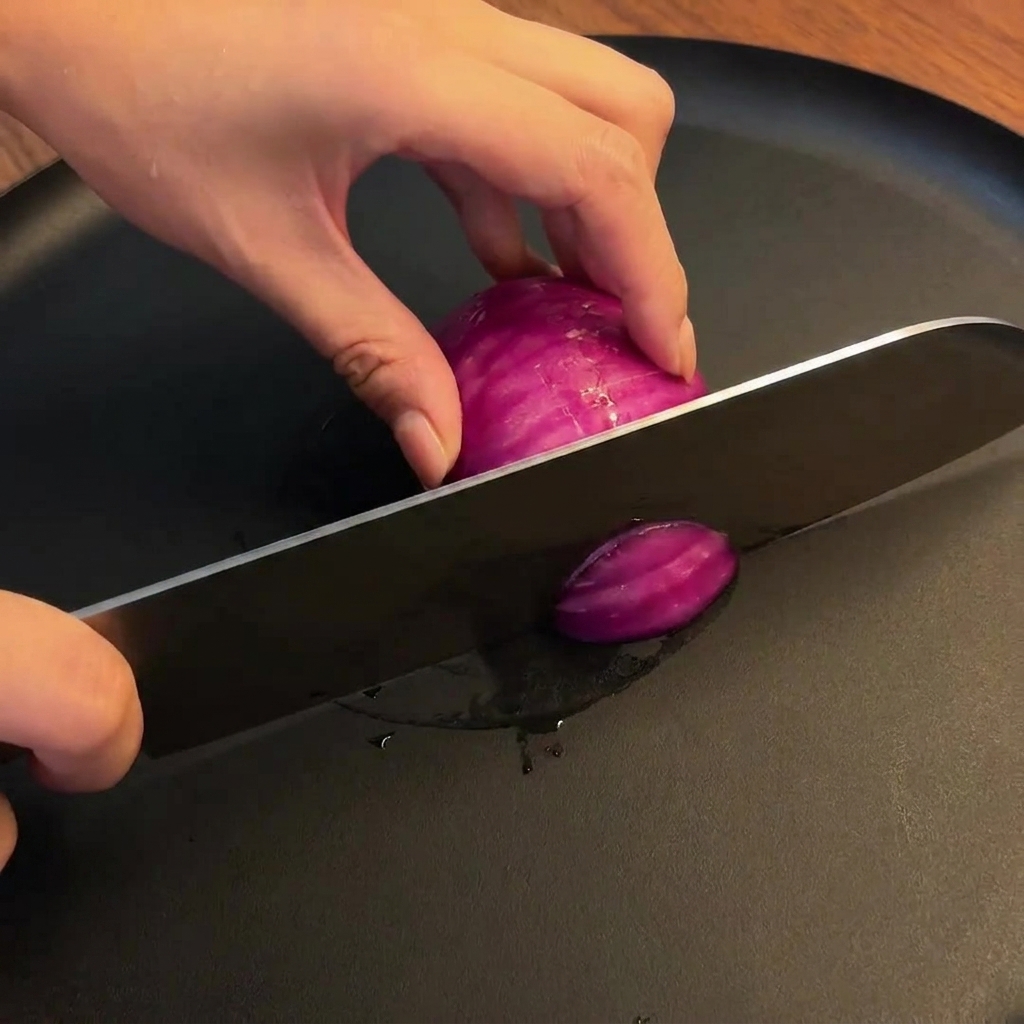 A red onion being sliced into thick, uniform pieces on a wooden cutting board with a chefs knife.