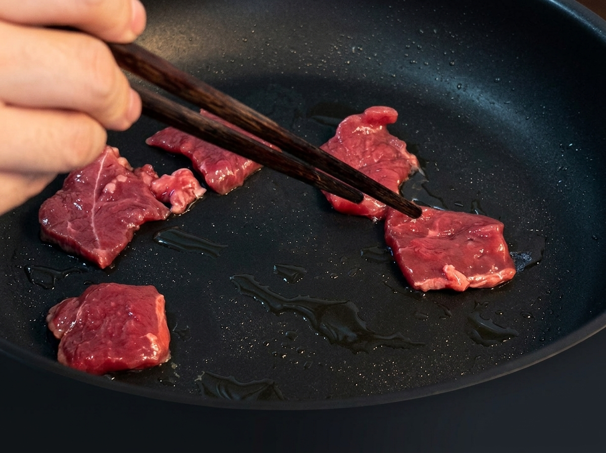 Thin slices of beef stir-frying in a hot, oiled pan, turning brown as they cook.