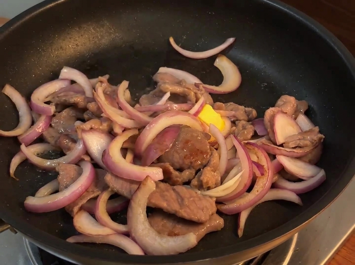 Stir-fry sauce being poured from a small bowl over the beef and onion mixture in a hot pan.