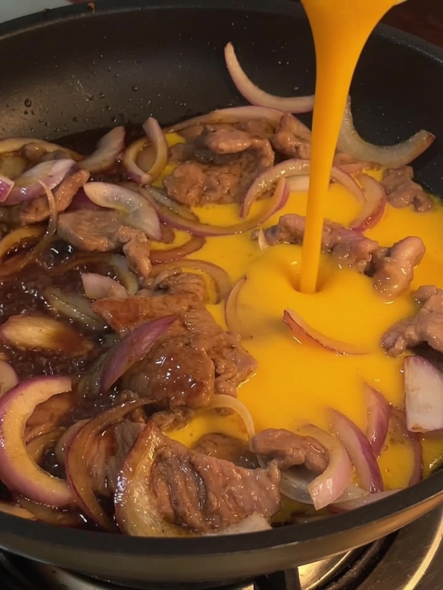 A close-up of whisked eggs being poured into a pan over stir-fried beef and onions.