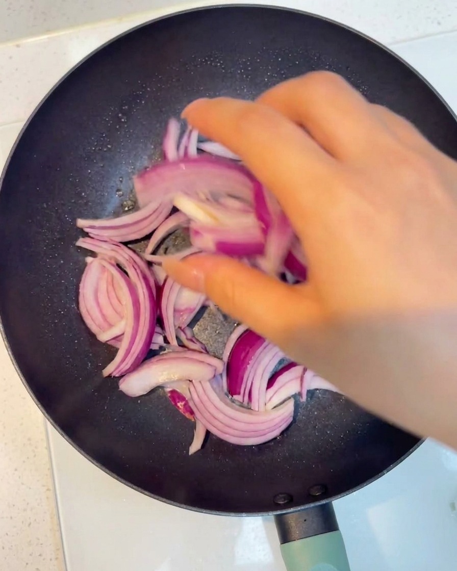 Slices of red onion being stir-fried in a hot pan until softened.