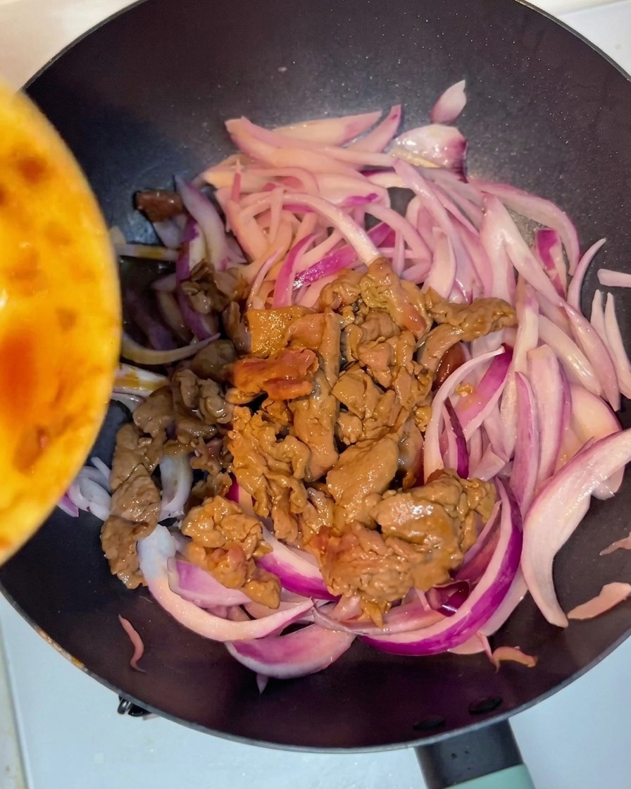 Cooked beef being added back into the pan with soft red onion slices, ready for mixing.