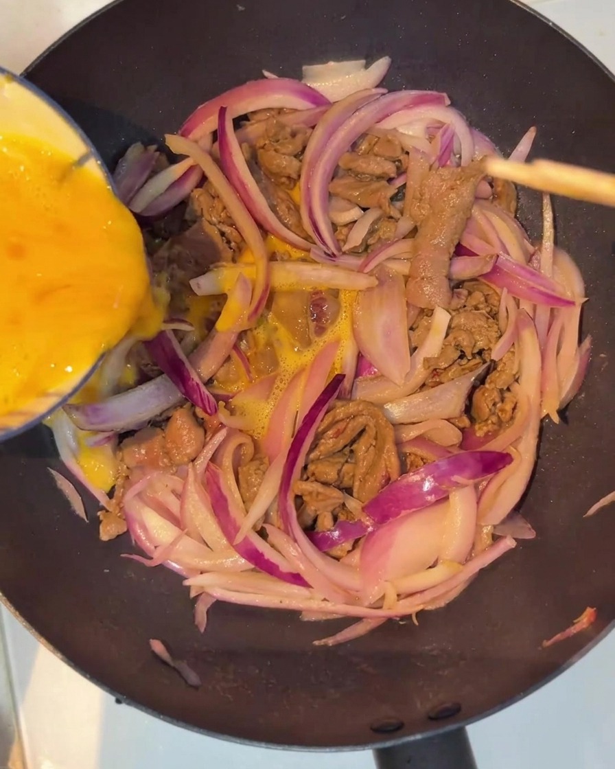 A mixture of beaten eggs being poured over the beef and onion base in the pan.