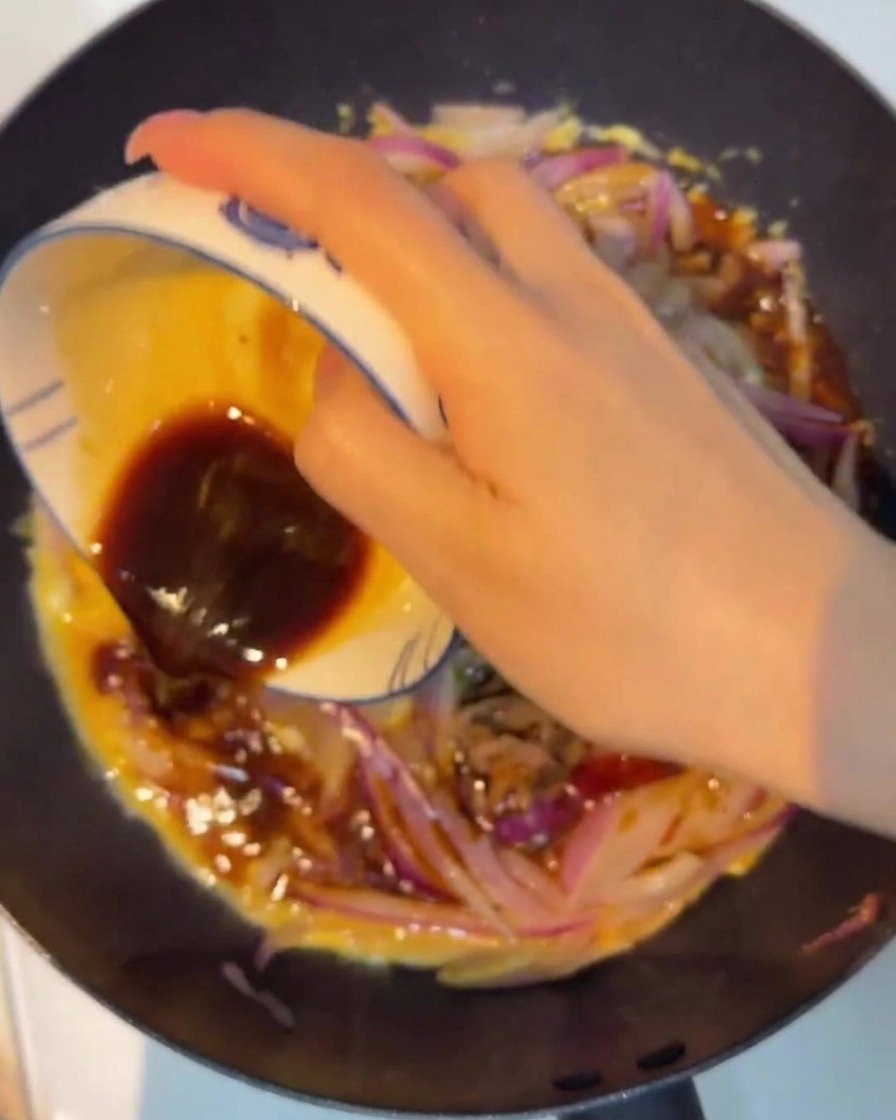 A dark, savory sauce being poured from a small bowl over the stir-fried beef, onions, and eggs in a non-stick pan.