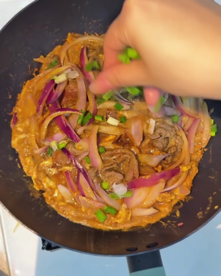 Freshly chopped green onions being sprinkled as a final garnish over the finished beef, onion, and scrambled egg stir-fry in the pan.