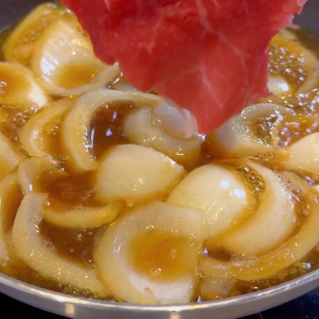 Thin slices of raw beef being placed into a pan with simmering onions and dark sauce.