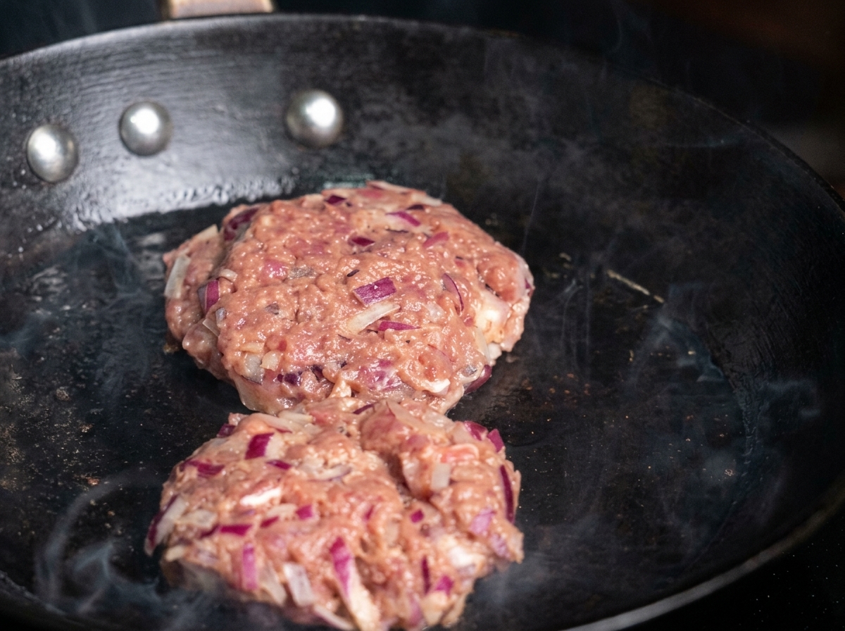 Two raw, seasoned beef patties being placed into a hot, oiled skillet, with wisps of smoke visible.