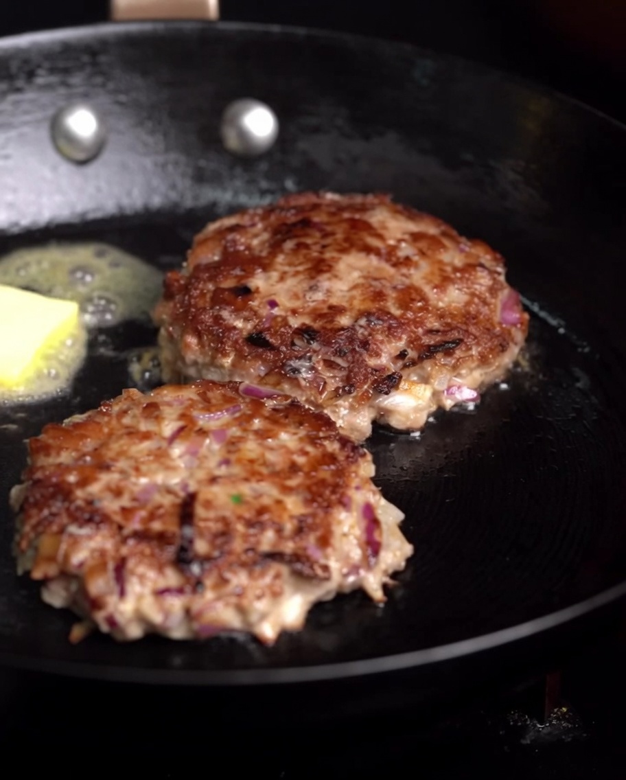 Beef patties frying in a pan with a melting cube of butter and a fresh sprig of rosemary.