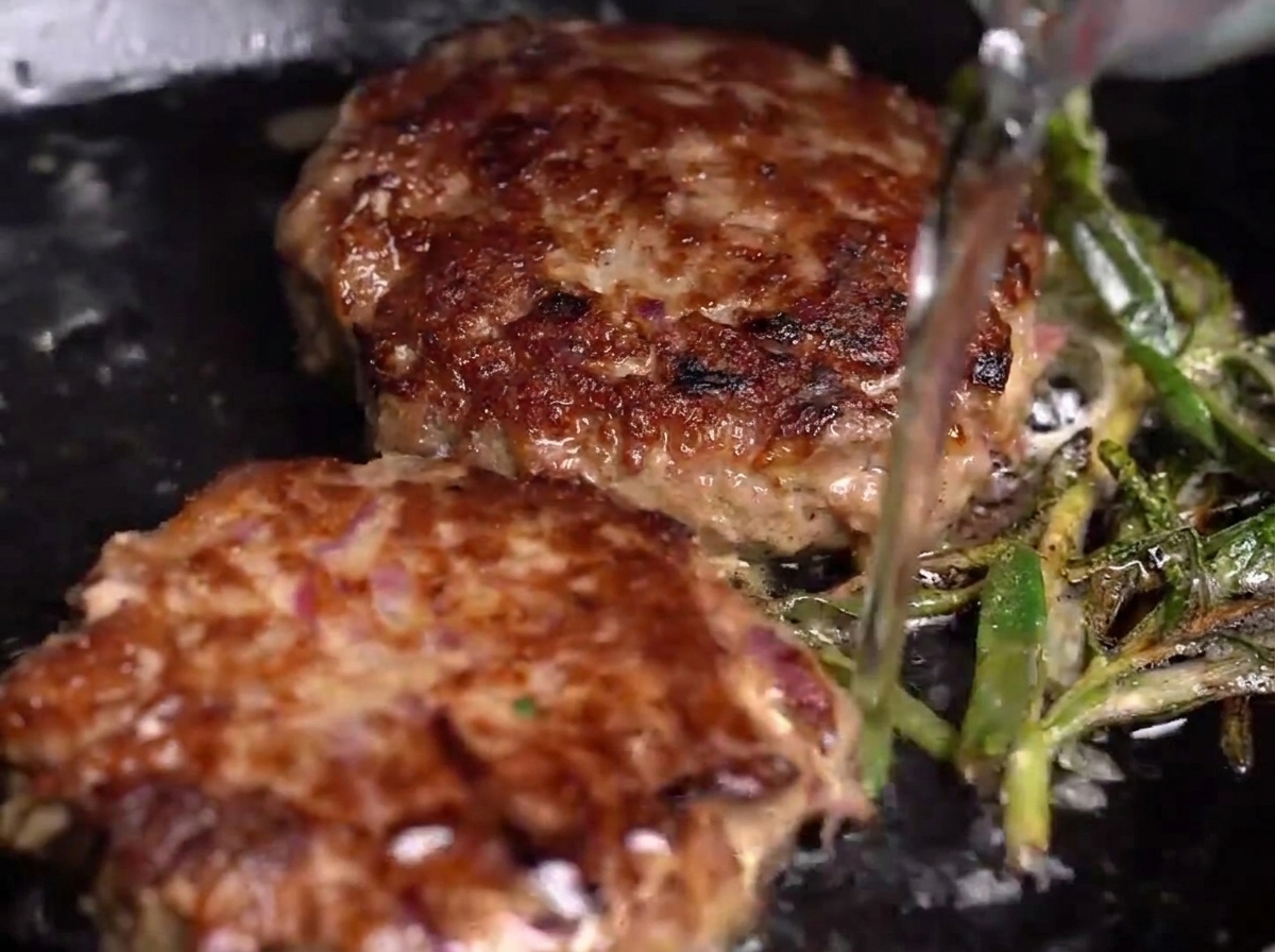 Water being poured into a hot pan containing seared beef patties and herbs, creating steam.