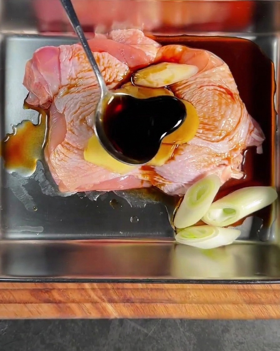 Soy sauce being poured from a spoon onto a raw chicken leg in a tray.