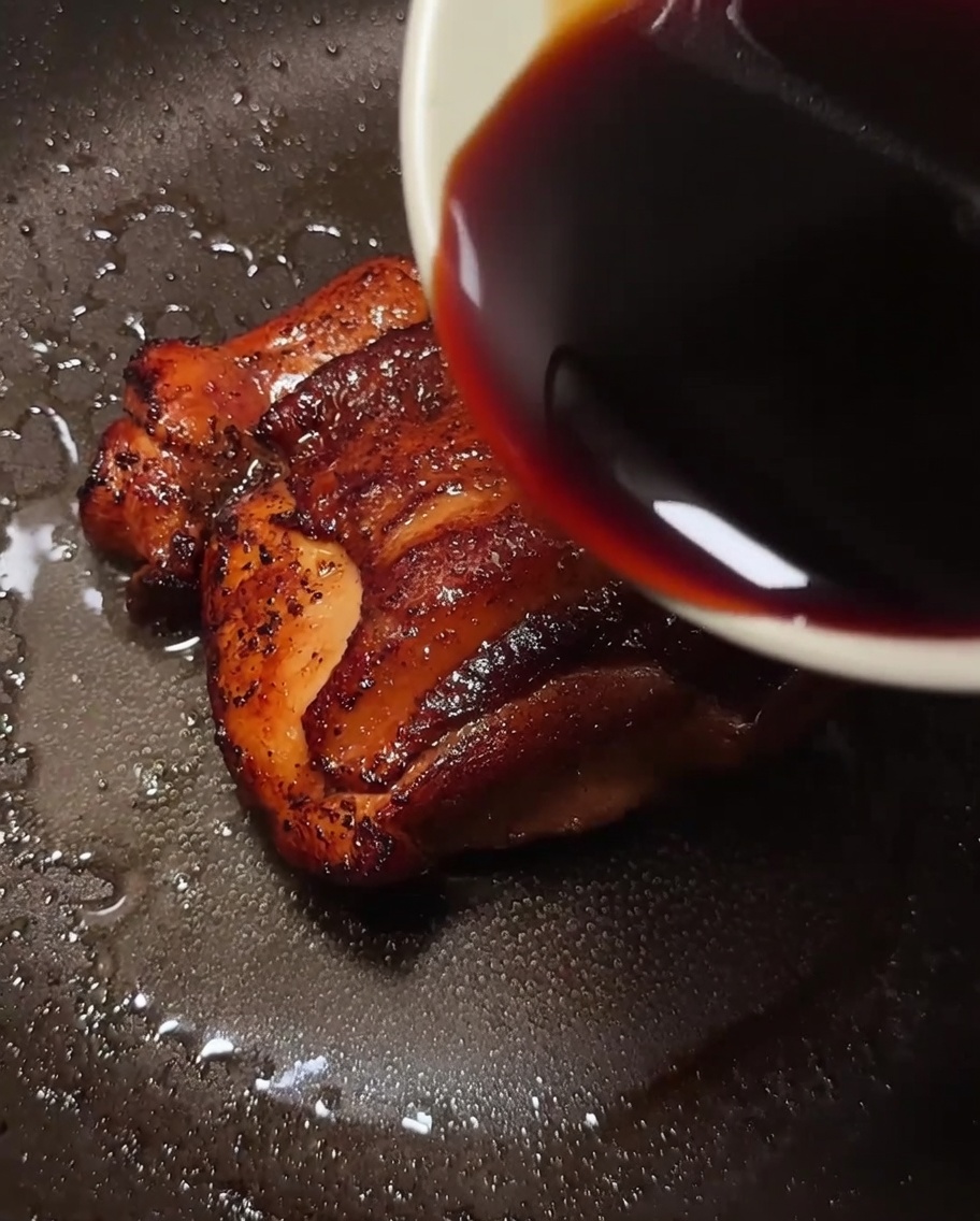 A pan containing a golden-brown chicken leg with a dark, savory sauce being poured over it.