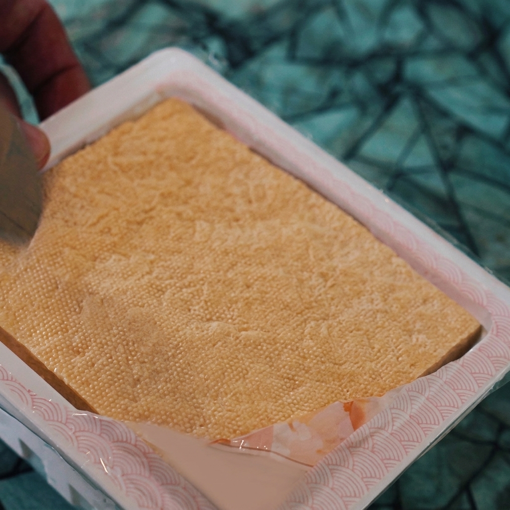 A hand using a knife to score a block of tofu into small squares inside its plastic packaging.