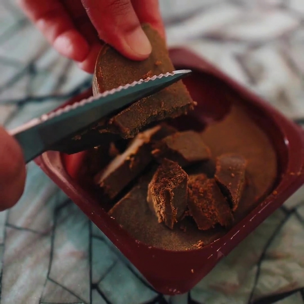 Hands using a serrated knife to chop solid blocks of brown Japanese curry into smaller pieces.