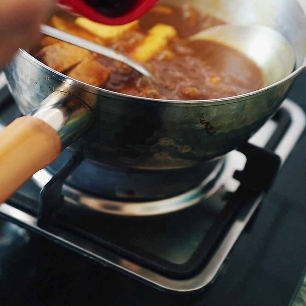 A spoonful of deep red Korean chili paste being poured into a pot of bubbling brown curry and tofu.