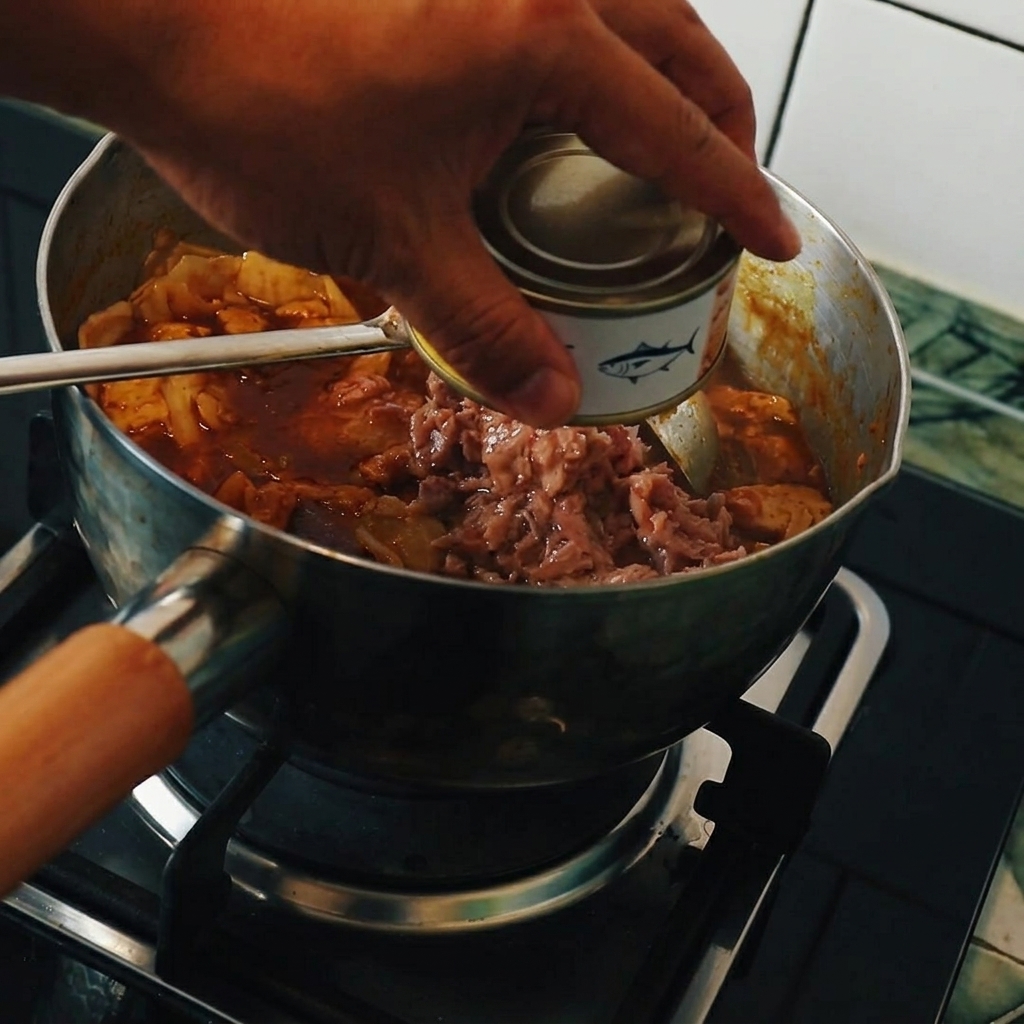 A hand holding a tuna can, pouring the contents into a pot filled with bubbling Japanese curry, tofu, and napa cabbage.