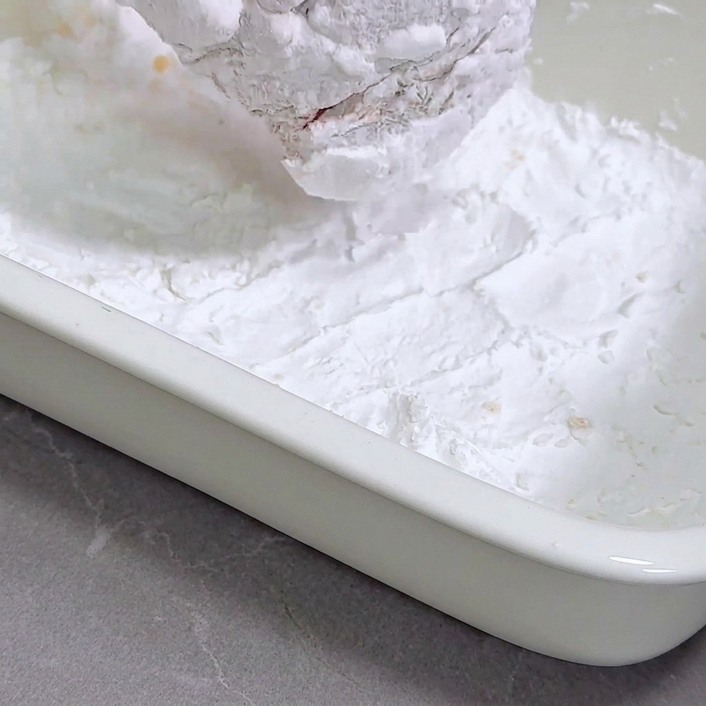 Marinated chicken breast being pressed into a white tray filled with cornstarch.