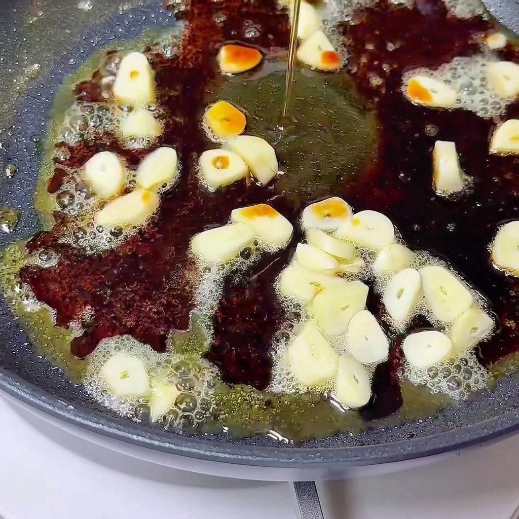Liquid being poured over sliced garlic and melted butter in a dark skillet.