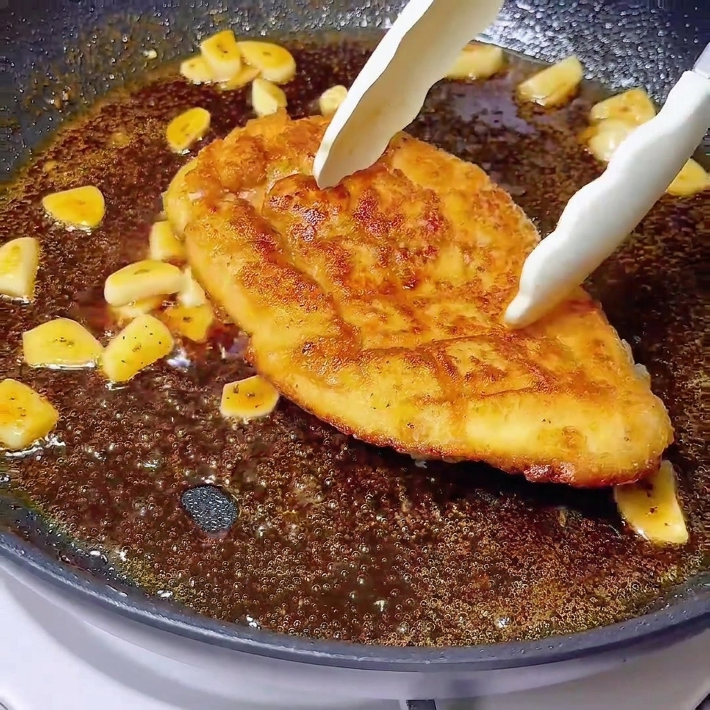 Tongs placing a golden-brown chicken breast into a bubbling dark glaze with sliced garlic.