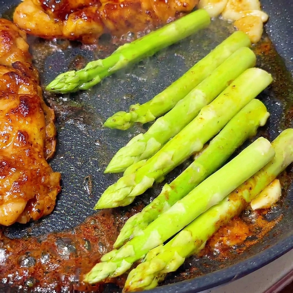 Fresh bright green asparagus spears simmering next to glazed chicken breasts in a skillet.