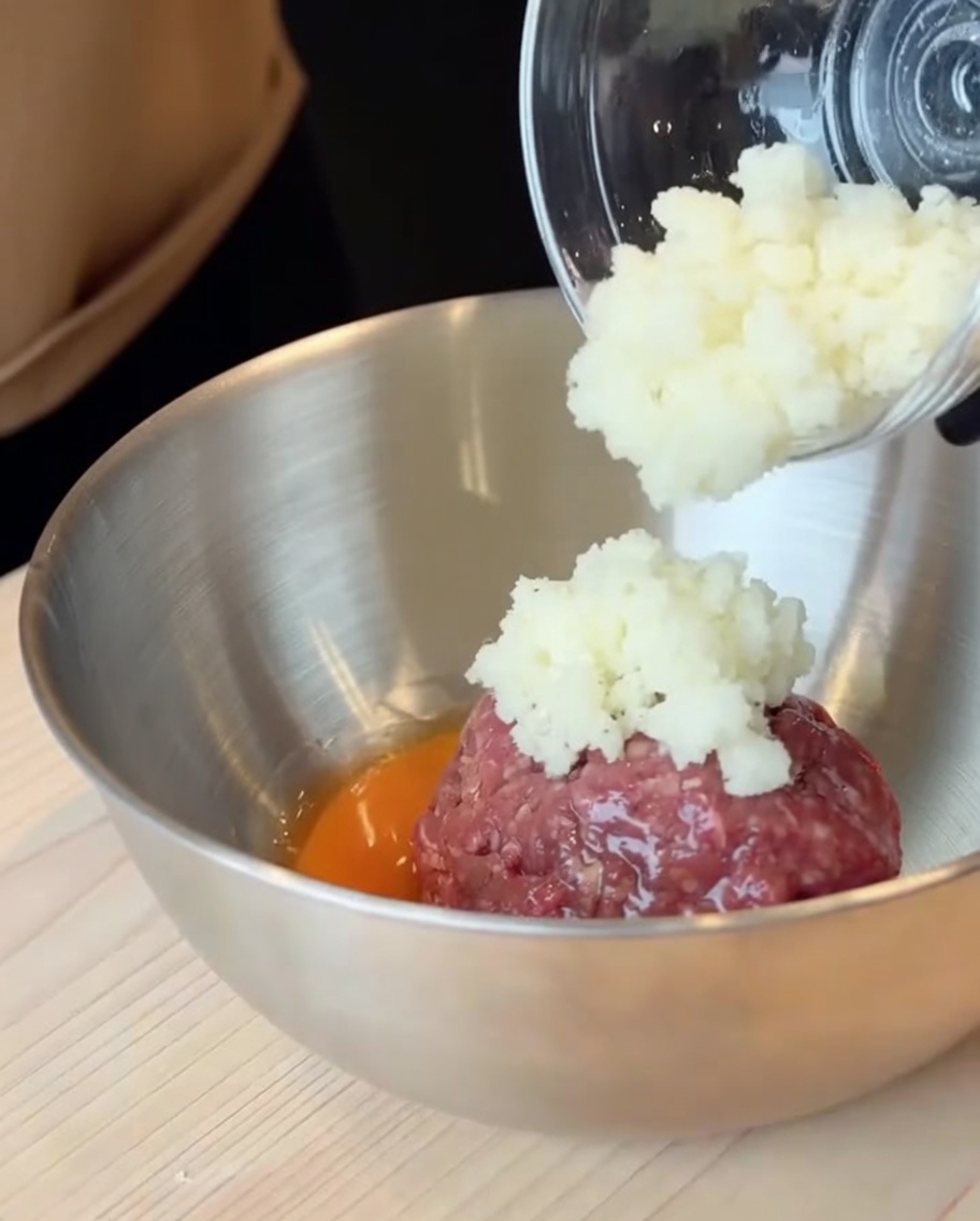 Raw ground beef and a cracked egg in a large stainless steel bowl with diced onions being added.