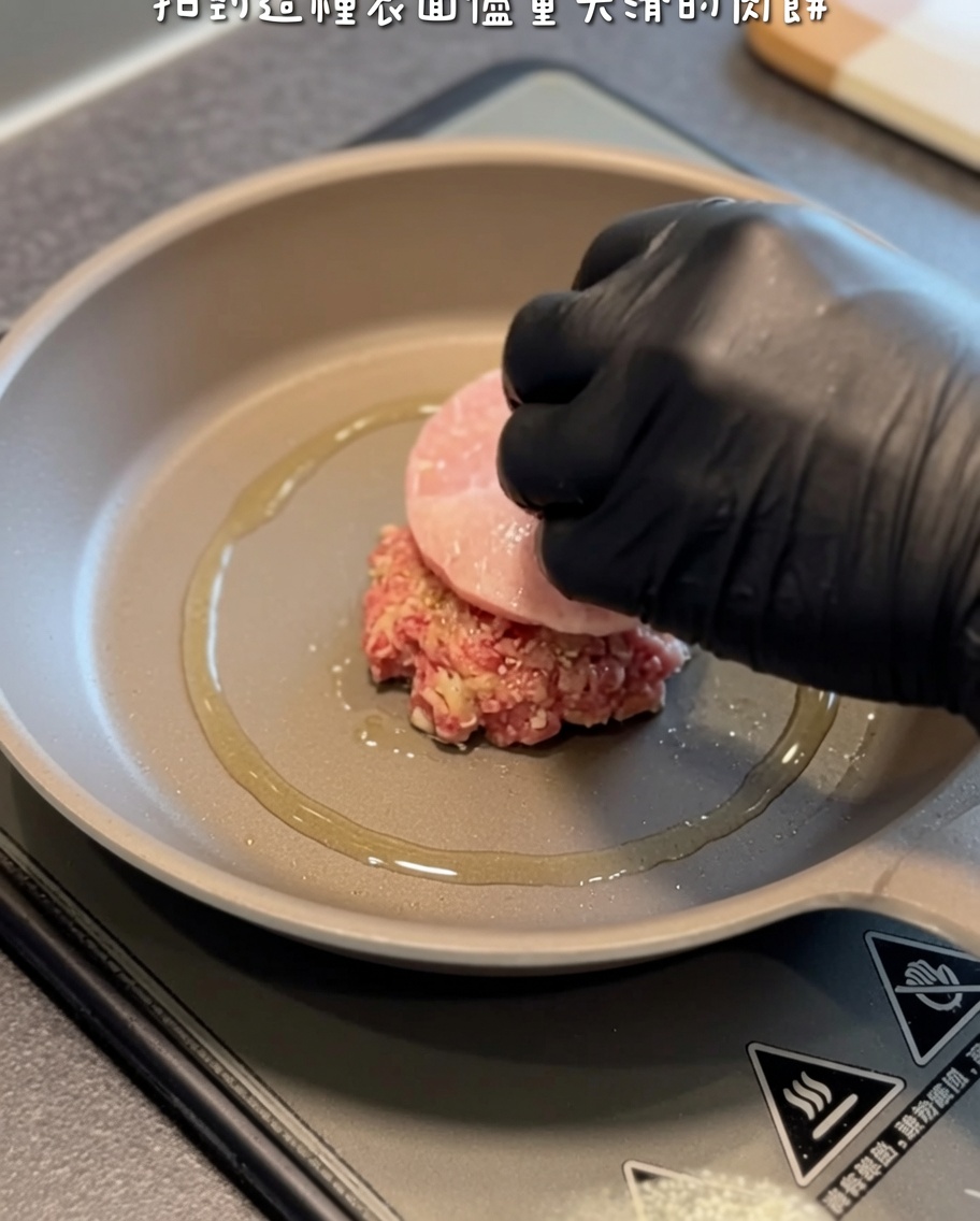 A raw hamburger patty being carefully lowered into a hot, oiled skillet using a black-gloved hand and spatula.