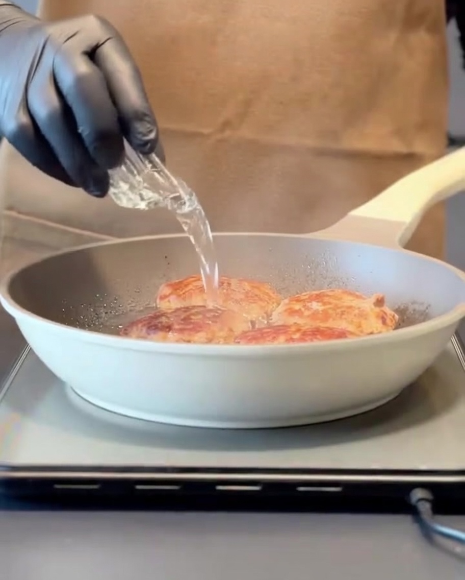 Water being poured into a hot pan containing seared meat patties, creating steam before covering with a lid.
