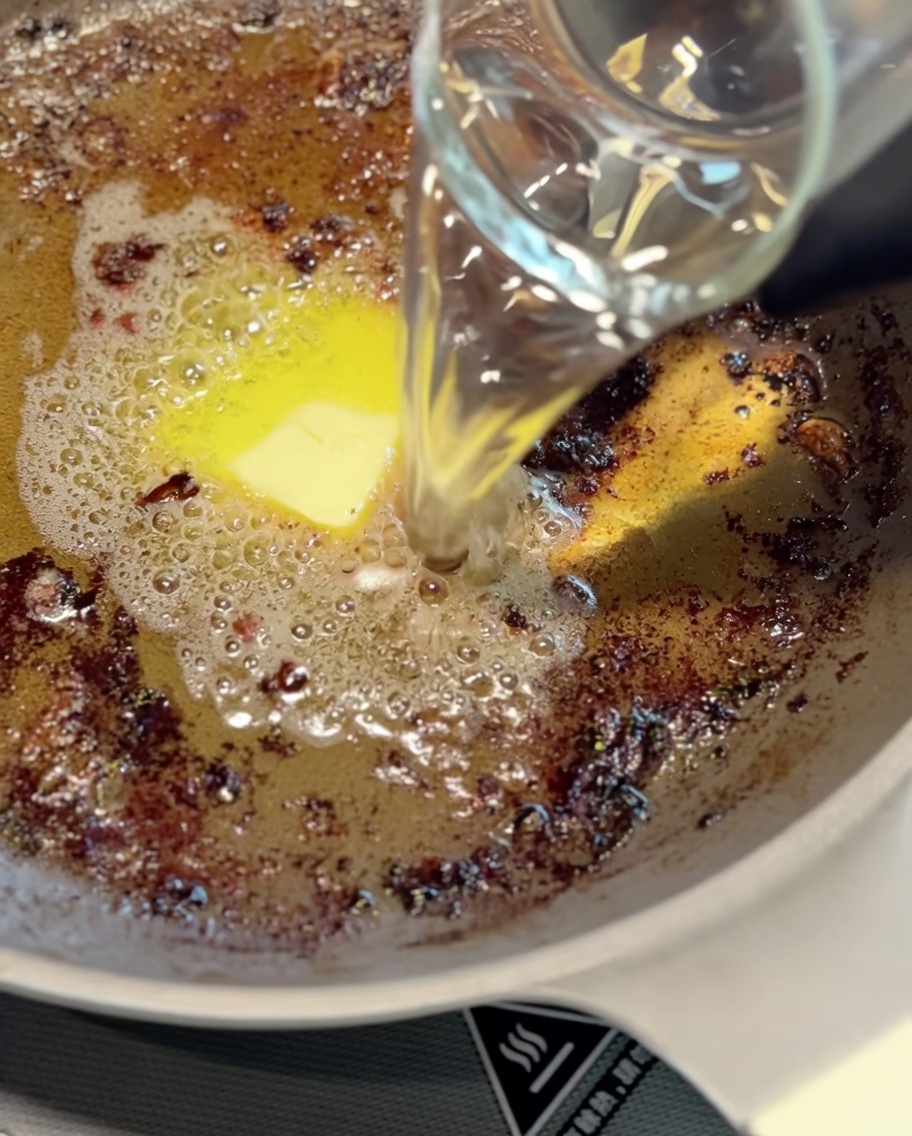 A metal ladle stirring browned pan juices and water in a skillet to form a base for the sauce.