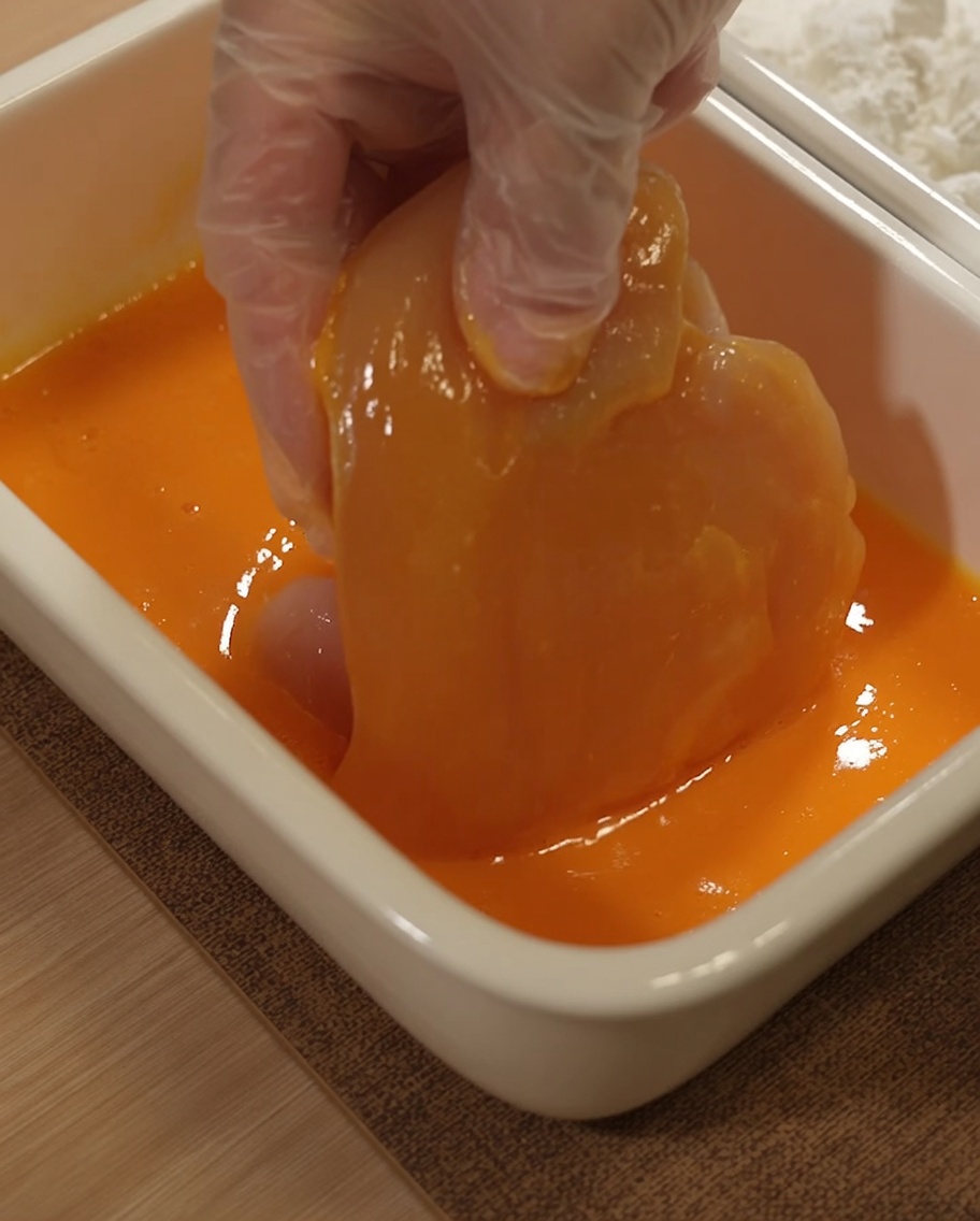 Chicken breast being submerged into a yellow egg wash mixture.