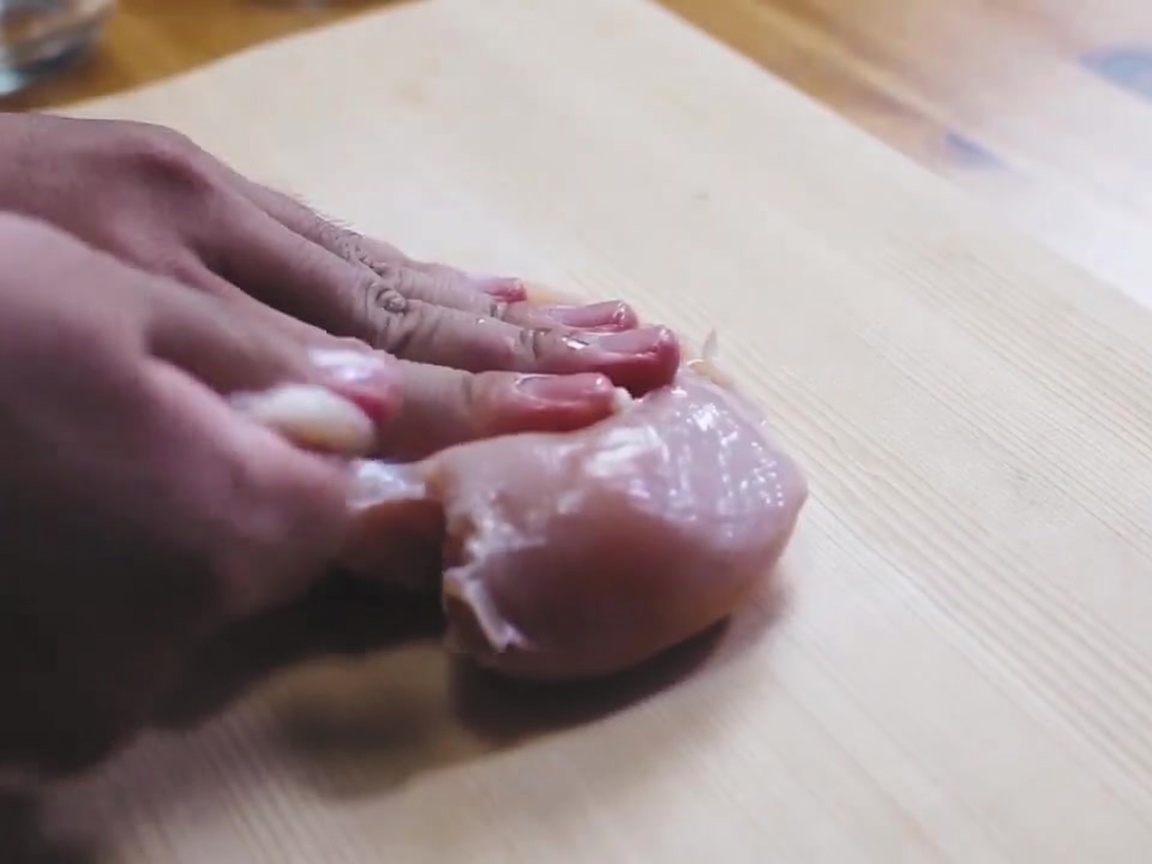 A raw chicken breast being sprinkled with salt on a wooden cutting board.
