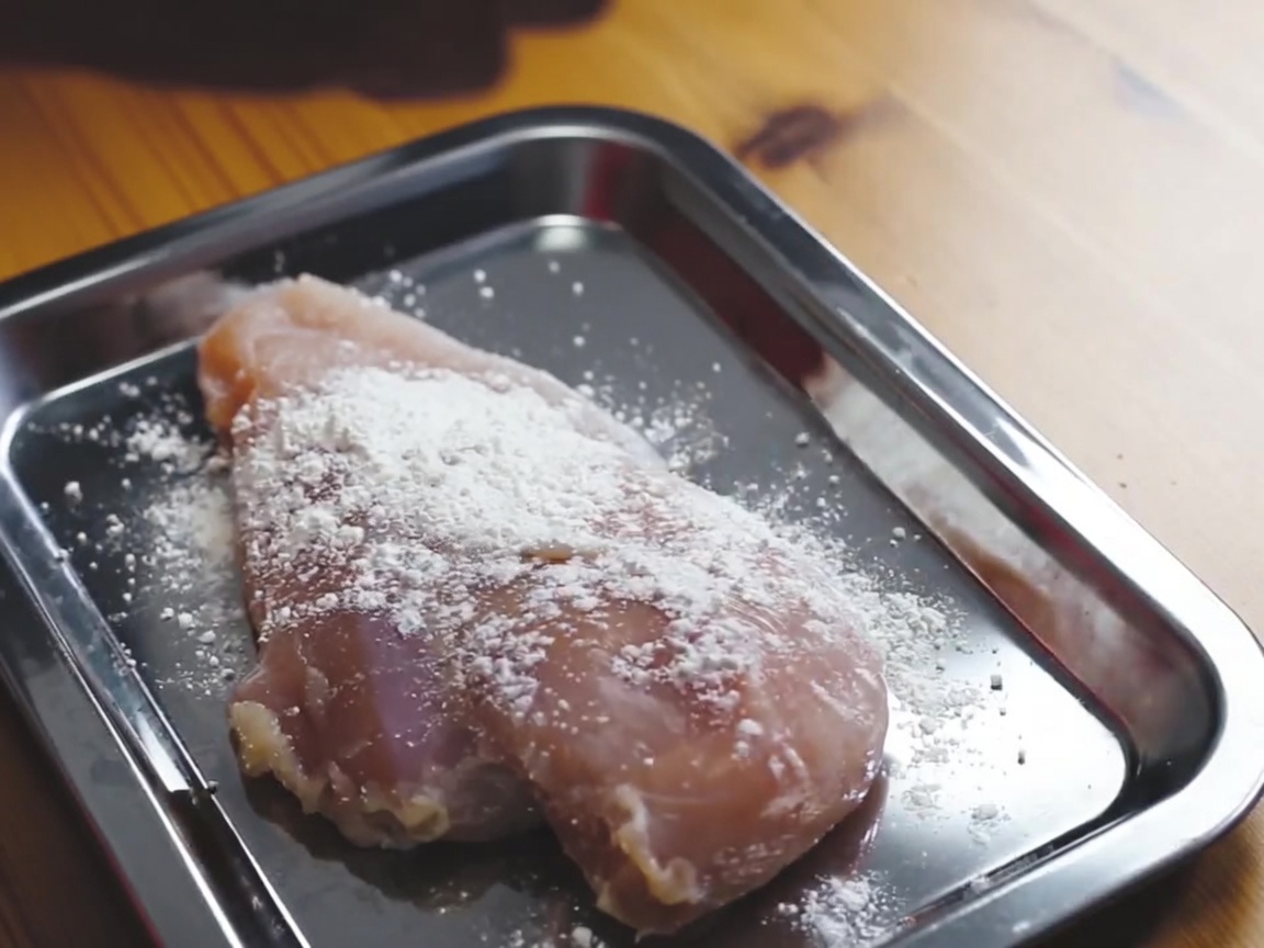 Raw chicken breast being coated in flour on a metal tray.