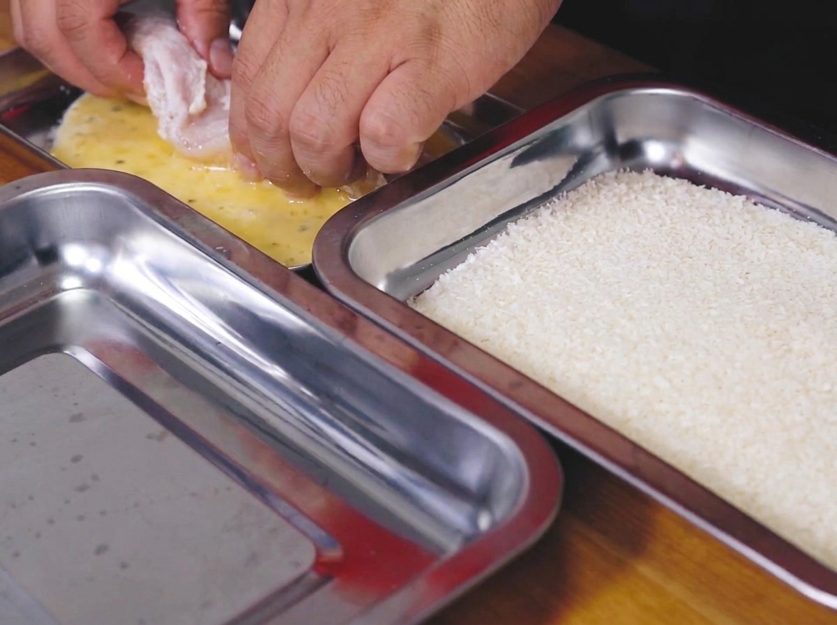Floured chicken breast being dipped into the egg and mayonnaise mixture in a tray.