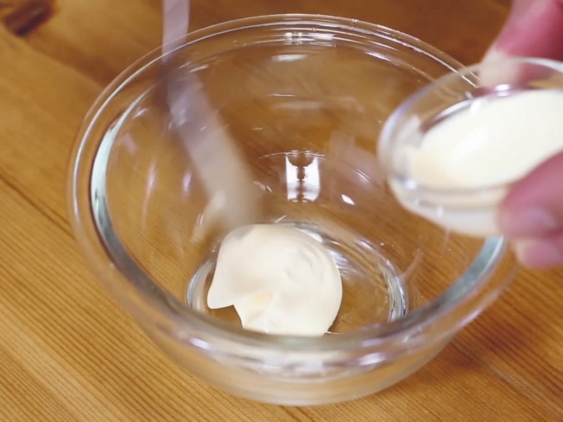 A small glass bowl containing a creamy mixture of mayonnaise and wasabi being stirred with a spoon.