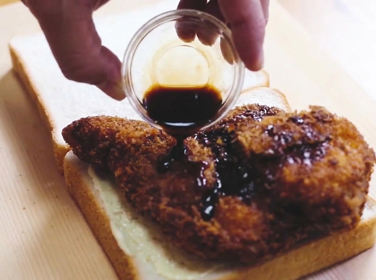 A breaded, golden-brown fried chicken cutlet being drizzled with dark sauce while resting on a piece of white bread.
