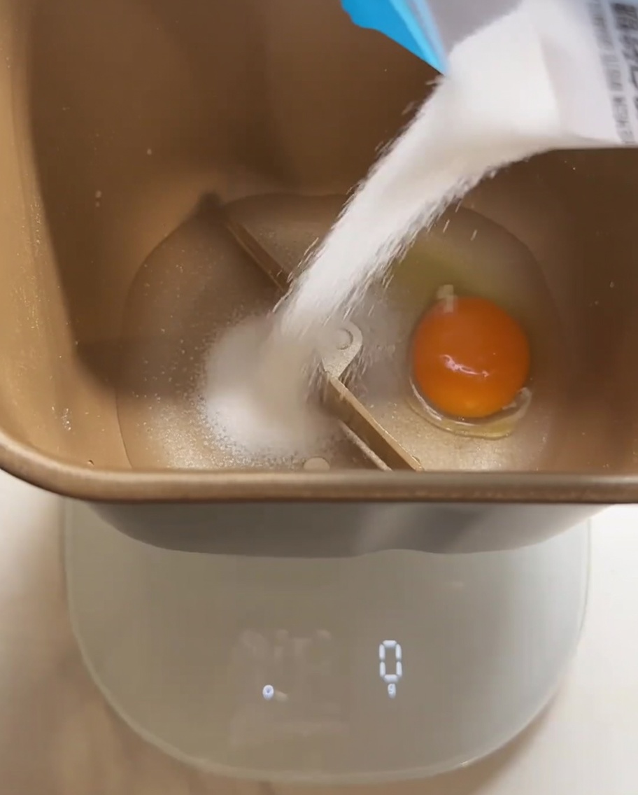 Ingredients for bread dough, including an egg and granulated sugar, being added to the bread machine container.