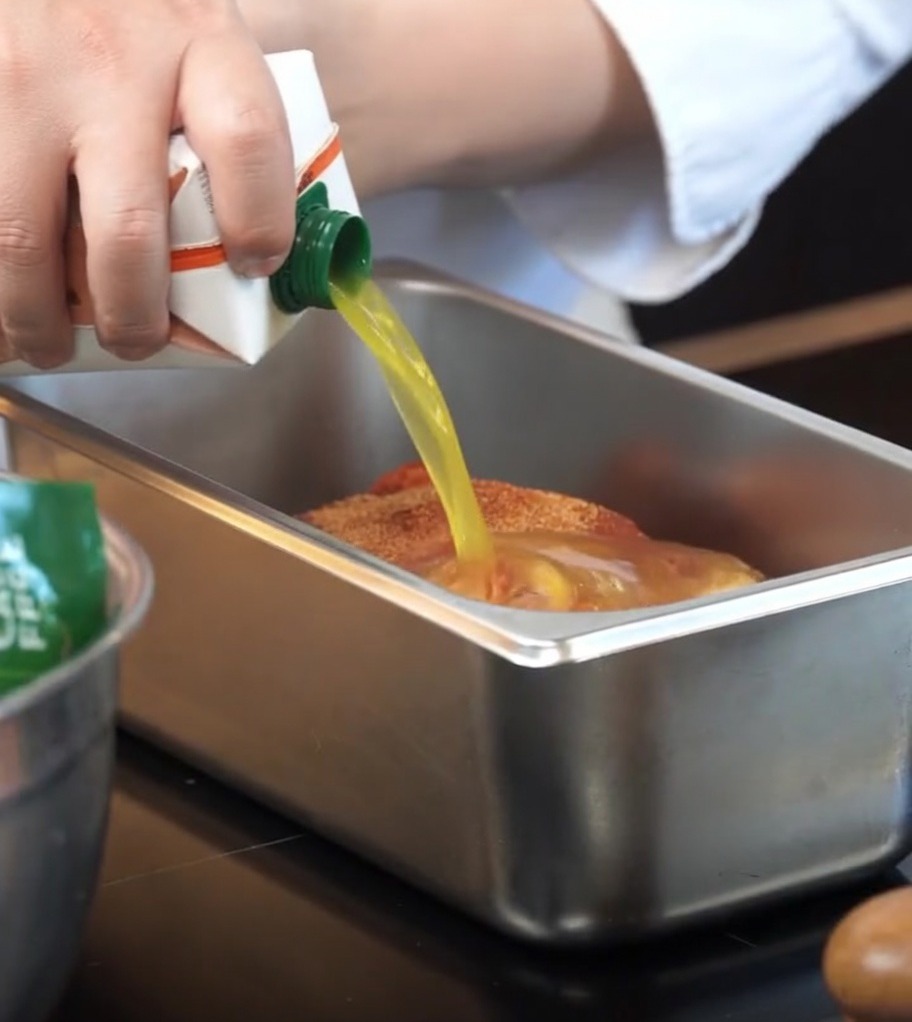 Orange juice being poured from a carton into a baking pan with seasoned pork.