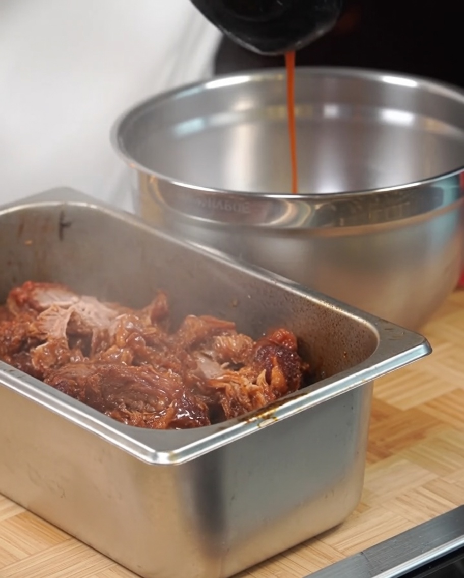 Shredded pork in a metal tray being seasoned with pan juices poured from a ladle.