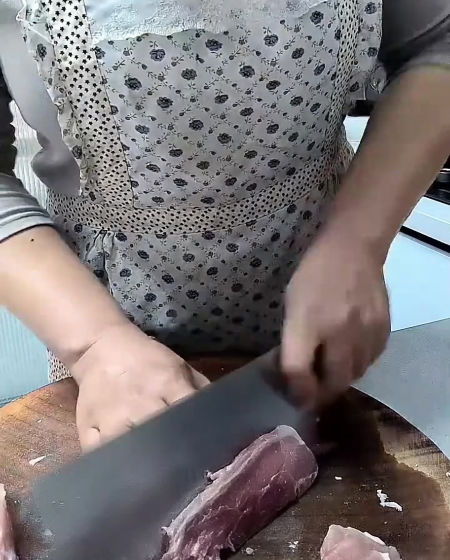 A sharp cleaver cutting through a piece of raw lean pork on a wooden cutting board.