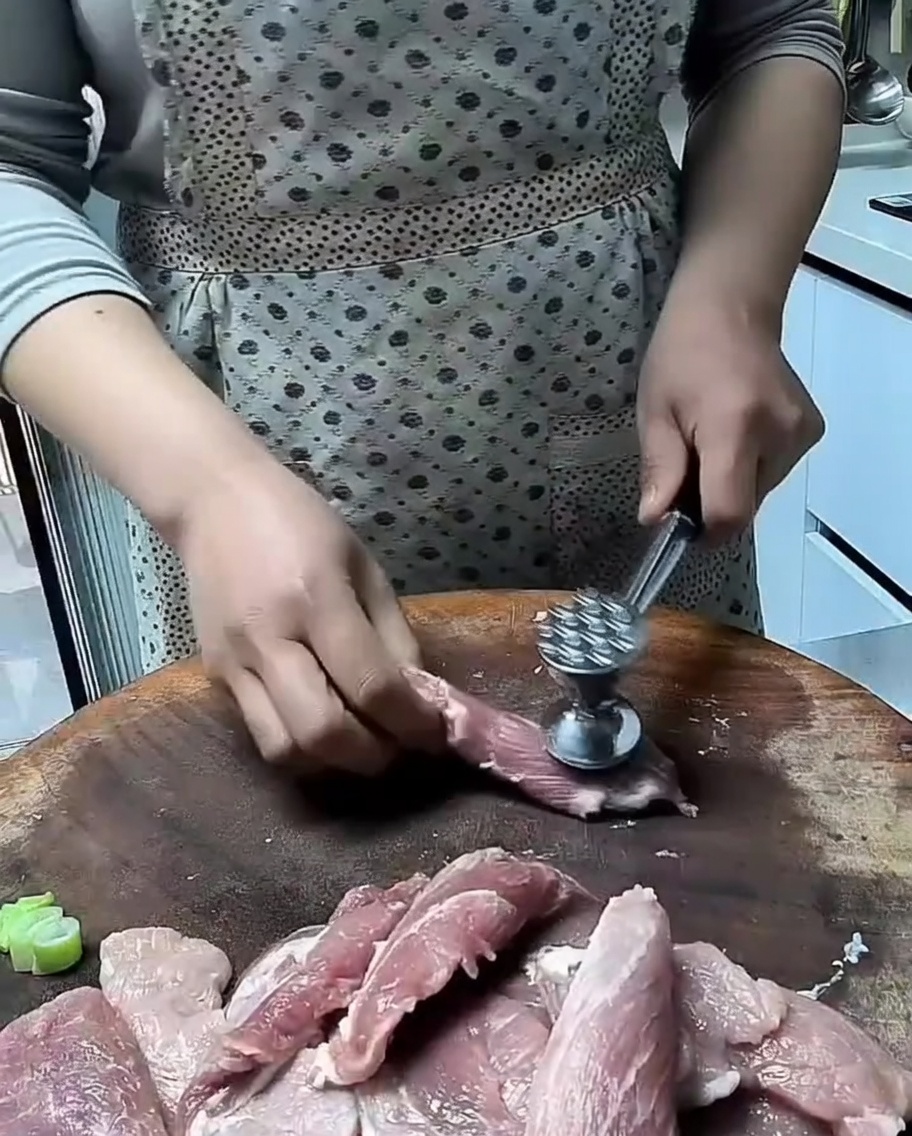 A metal meat mallet tenderizing thin slices of raw pork on a wooden surface.