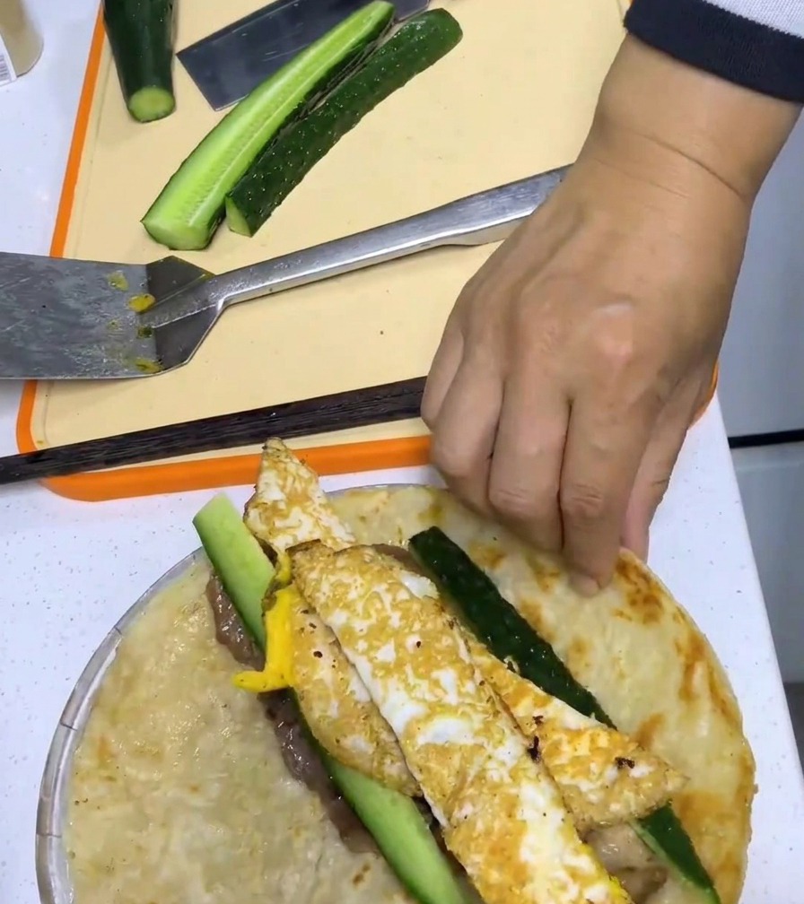 A hand rolling a scallion pancake tightly around pork, egg, and cucumber fillings.