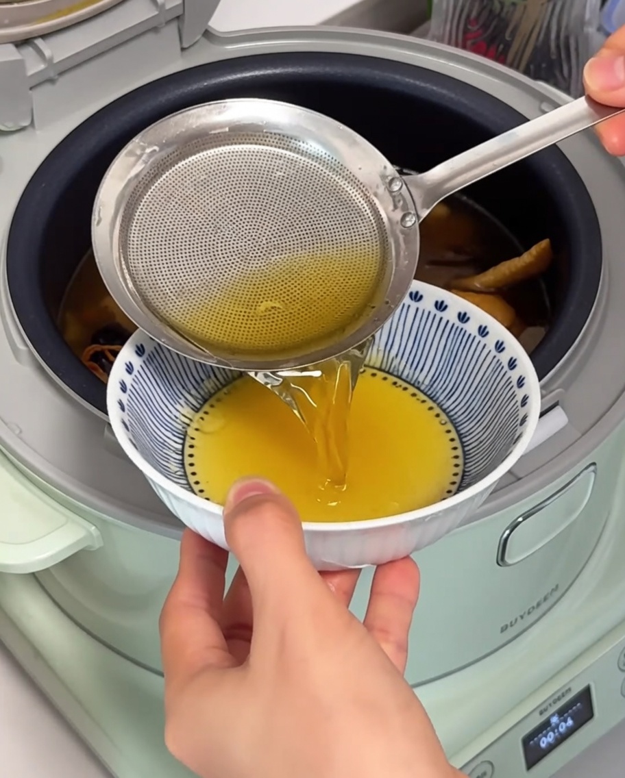 A close-up of a fine-mesh strainer lifting clear, golden chicken oil off the top of the simmering soup in an electric cooker.