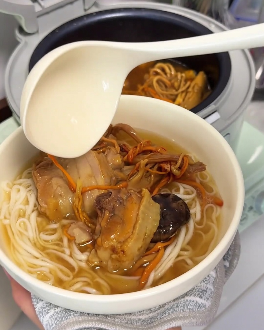 A large ladle pouring rich, golden chicken and mushroom soup over a bowl of freshly cooked noodles.