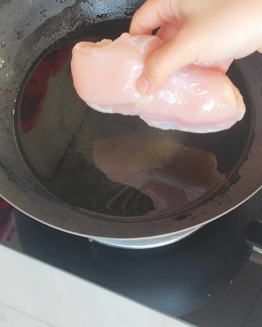 A chicken breast submerged in boiling water inside a wok alongside aromatics like ginger and green onion.